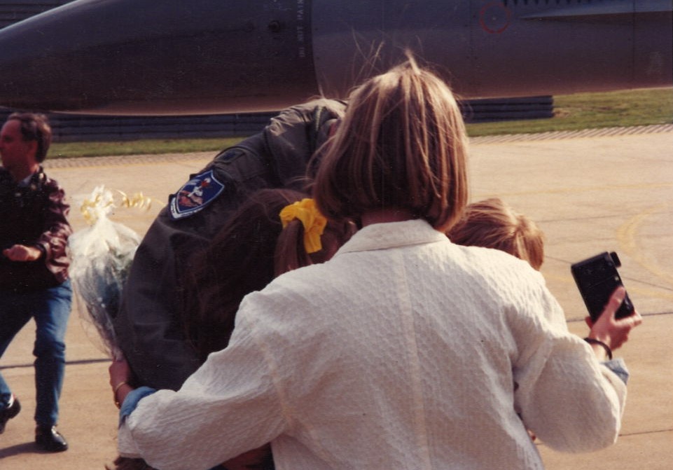 Spring 1991 — flight suit, flowers, a Polaroid in someone's hand, and three small arms holding on.